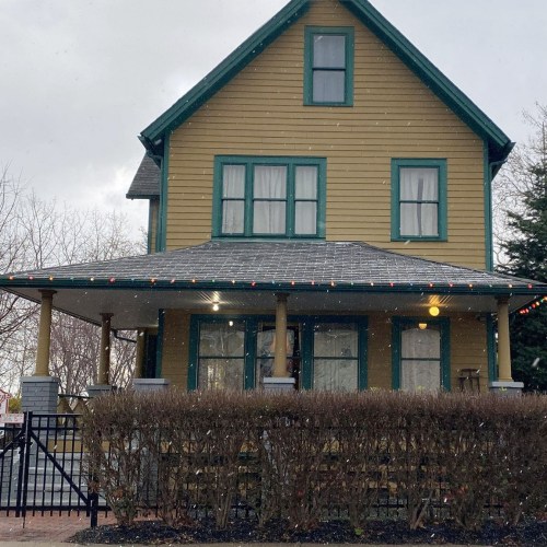 A yellow house with green trim, snow falling, and holiday lights on the porch, surrounded by bare trees.