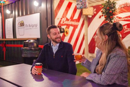 A man and woman sit at a table, smiling and enjoying drinks in a festive outdoor setting with holiday decorations.