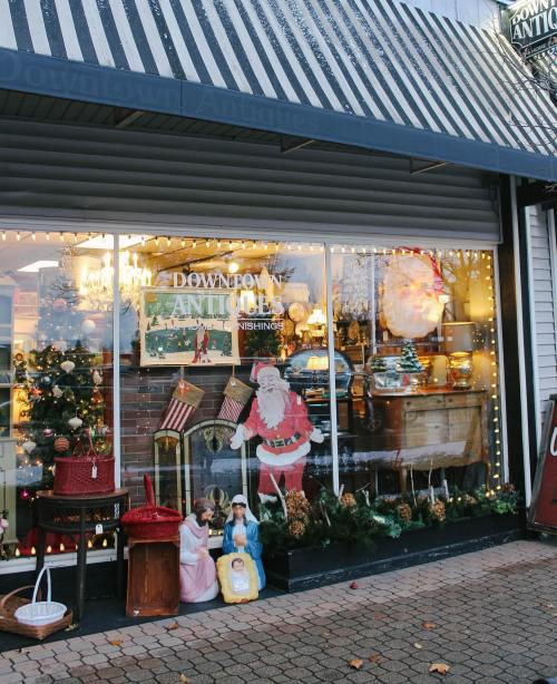 A festive antique shop window decorated for the holidays, featuring Santa, dolls, and various vintage items.