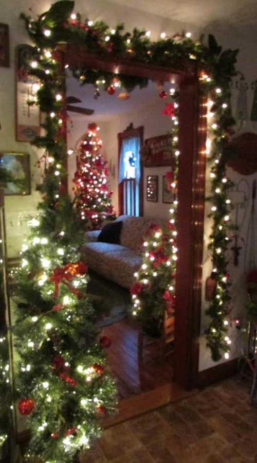 A warmly decorated doorway with garlands and lights, leading to a cozy room with a Christmas tree in the background.