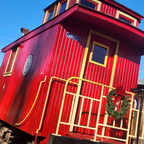 A bright red train caboose with yellow trim, featuring a wreath on the railing, set against a clear blue sky.