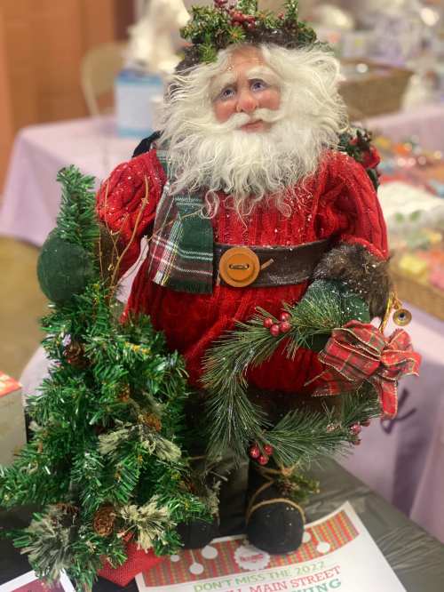 A festive Santa figure in a red outfit, holding a small tree and a gift, decorated with greenery and a plaid scarf.