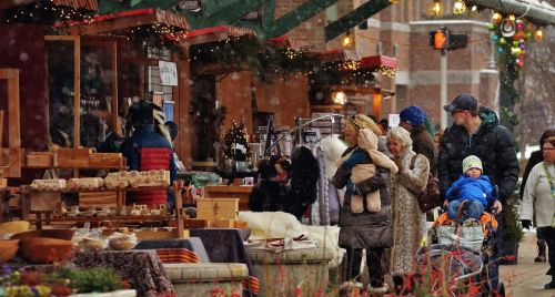 A bustling outdoor market scene with shoppers browsing stalls, festive decorations, and a light snowfall.