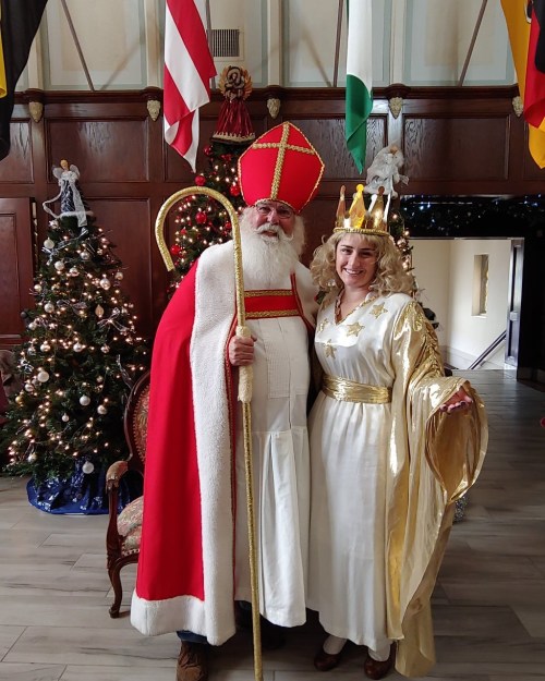 A man in a red bishop's outfit and a woman in a white gown and crown pose together in a festive setting.