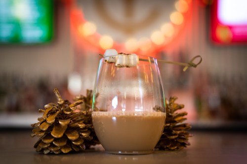 A glass of creamy cocktail garnished with a marshmallow skewer, surrounded by pinecones, with a blurred bar background.