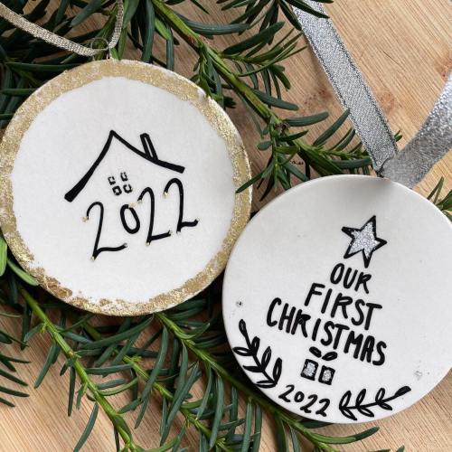 Two decorative Christmas ornaments on greenery, one reads "2022" and the other "Our First Christmas 2022."