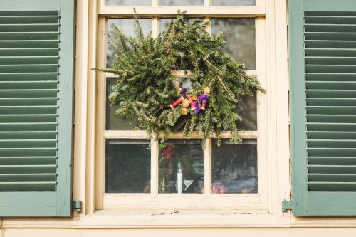 A festive wreath made of greenery and colorful ribbons hangs on a window with green shutters.