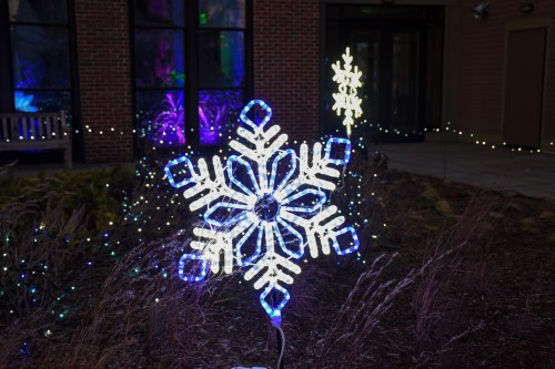 A large illuminated snowflake decoration in blue and white, surrounded by colorful lights in a garden setting.