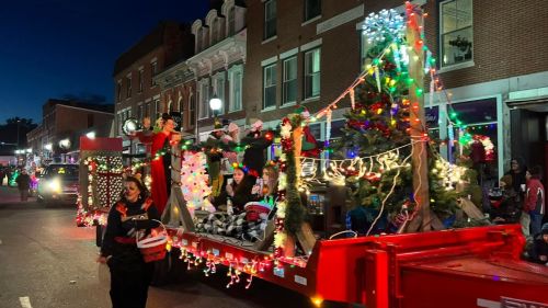 A festive parade float decorated with lights, a Christmas tree, and people in holiday attire, set against a twilight backdrop.
