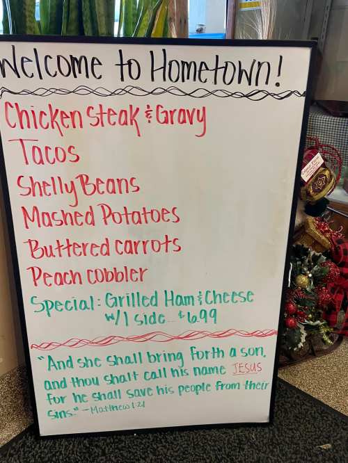 A menu board listing food items: chicken steak, tacos, sides, and a special for grilled ham and cheese. Bible verse included.