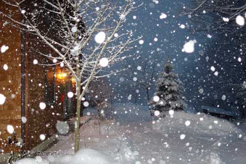 A snowy scene with falling snowflakes, a cozy house illuminated by warm light, and trees blanketed in white.