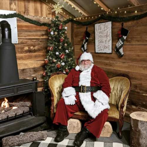 Santa Claus sits in a cozy room by a fireplace, with a decorated Christmas tree and stockings hanging nearby.