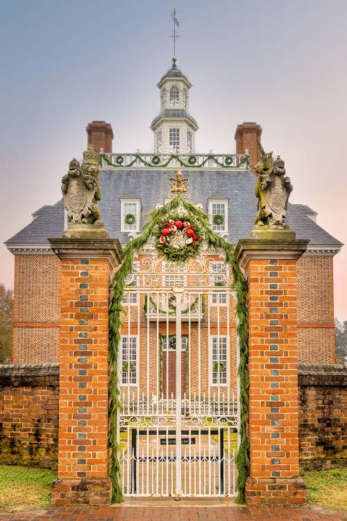 A grand brick gate adorned with greenery leads to a historic mansion with a distinctive cupola and weather vane.