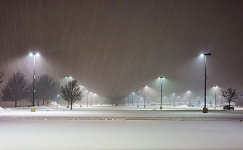 A snow-covered empty parking lot at night, illuminated by streetlights in a heavy snowfall.