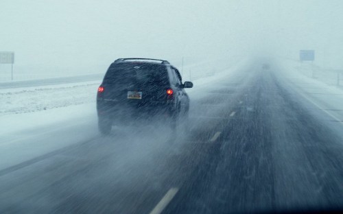 A black SUV drives through heavy snow, with visibility significantly reduced on a snowy road.