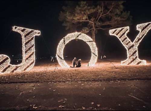 A couple sits beneath large illuminated letters spelling "JOY" in a dark park, surrounded by festive lights.