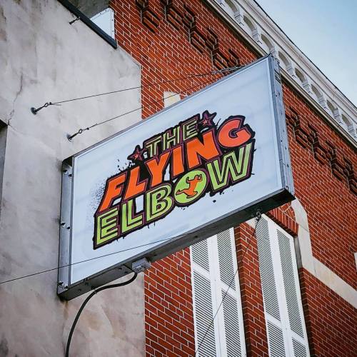 Sign for "The Flying Elbow" featuring bold, colorful lettering against a brick building backdrop.