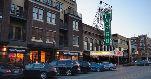 Historic Fargo Theatre sign illuminated at dusk, surrounded by buildings and parked cars on a city street.
