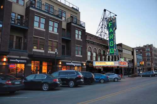 Historic Fargo theater with a neon sign, surrounded by brick buildings and parked cars on a quiet street at dusk.