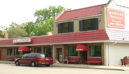 A restaurant with a red roof and awnings, featuring outdoor seating and a sign that reads "Homer's Restaurant."