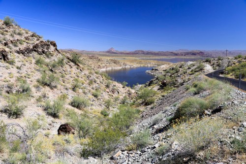 A scenic view of a desert landscape with a winding river, rocky hills, and sparse vegetation under a clear blue sky.