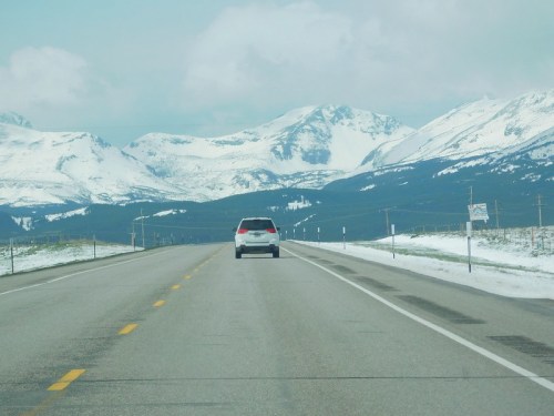 A car drives on a highway with snow-capped mountains in the background under a cloudy sky.