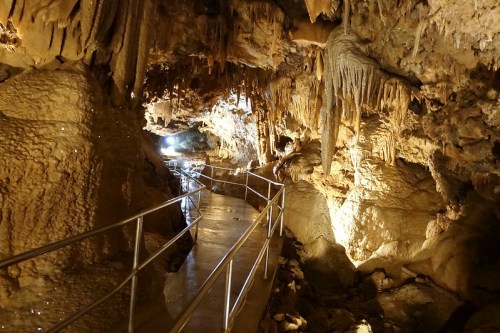 A dimly lit cave interior with a metal walkway, featuring stalactites and stalagmites along the rocky walls.
