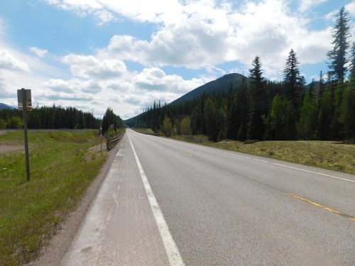 A wide, empty road stretches through a forested landscape under a partly cloudy sky. Mountains rise in the distance.