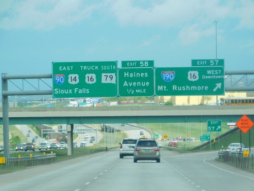 Highway sign indicating exits for Sioux Falls, Haines Avenue, and Mt. Rushmore, with traffic on the road below.