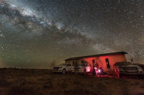 A house under a starry sky with the Milky Way, surrounded by parked vehicles and a person setting up equipment.