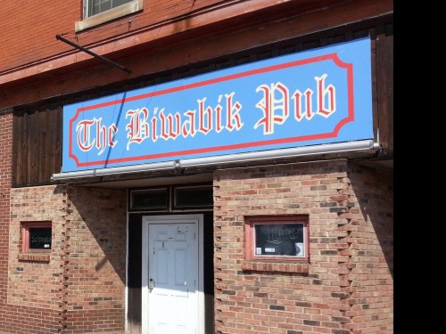 Sign for "The Biwabik Pub" on a brick building, featuring blue background and decorative red border.