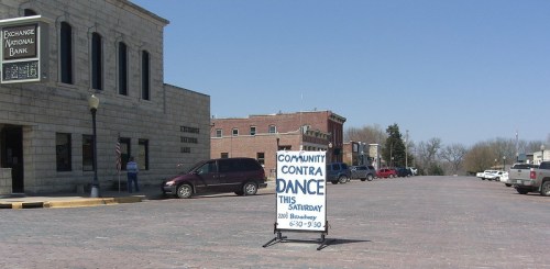 A street scene featuring a sign for a community contra dance, with buildings and parked cars in the background.