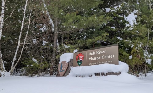 Sign for Ash River Visitor Center at Voyageurs National Park, surrounded by snow and trees.