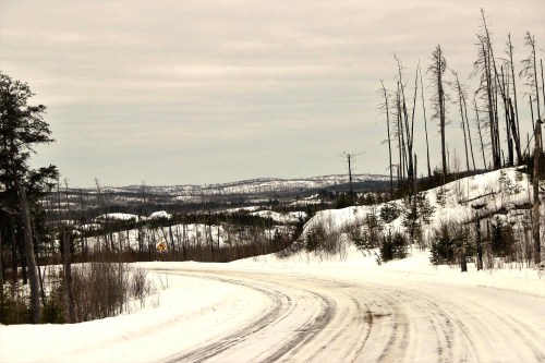Winding snowy road through a winter landscape with bare trees and distant hills under a cloudy sky.