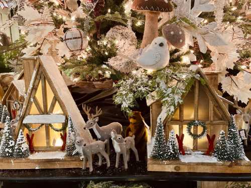 Decorative winter scene with two wooden houses, reindeer figurines, and festive greenery under a Christmas tree.