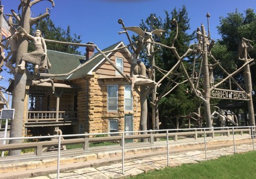 A whimsical house with tree sculptures and figures, featuring a sign that reads "Garden of Eden." Bright blue sky above.