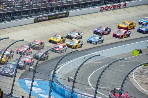 Aerial view of multiple NASCAR cars racing on a track at Dover, with grandstands in the background.