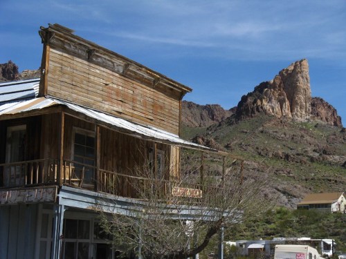 A weathered wooden building in the foreground with a rocky mountain peak in the background under a clear blue sky.