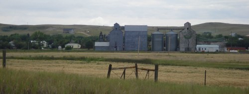 Rural landscape featuring silos and farm buildings against a backdrop of rolling hills and a cloudy sky.
