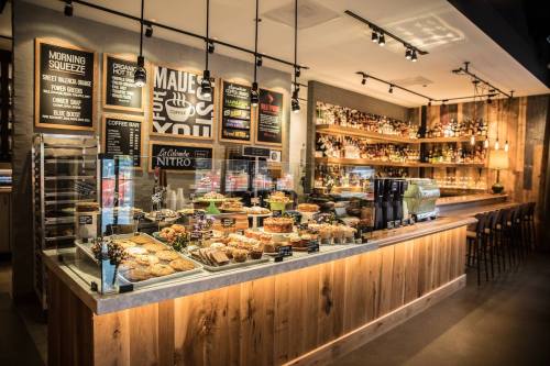 A modern café interior featuring a display of pastries, a coffee bar, and a wooden counter with shelves of drinks.