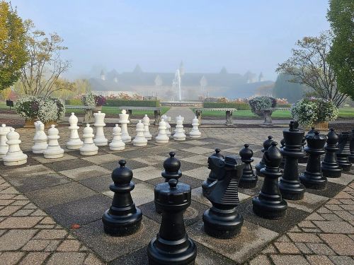 A large outdoor chessboard with black and white pieces, surrounded by greenery and a misty background.