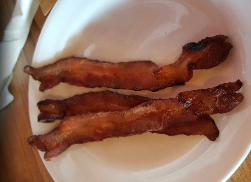 Three crispy strips of bacon on a white plate, resting on a wooden table.