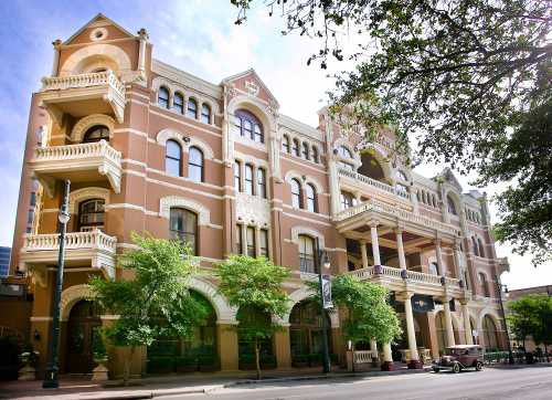 Historic building with ornate architecture, balconies, and lush greenery in front, set against a clear blue sky.