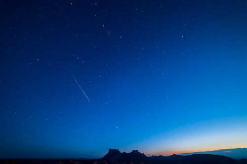 A starry night sky with a faint meteor streaking across, silhouetted mountains on the horizon against a gradient blue to orange.