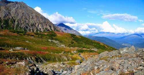 A scenic mountain landscape with rocky terrain, lush greenery, and a clear blue sky dotted with clouds.