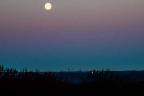 A full moon rises over a distant city skyline, with a gradient sky transitioning from dusk to night.