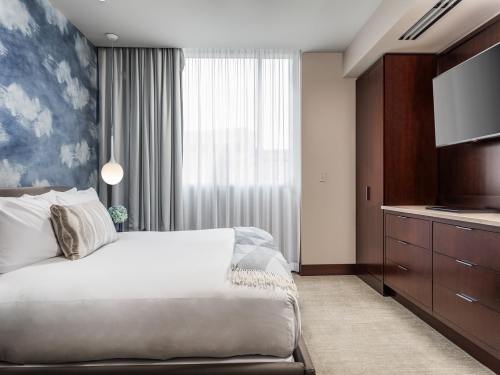 A modern hotel room featuring a bed, wooden furniture, a TV, and large windows with sheer curtains.