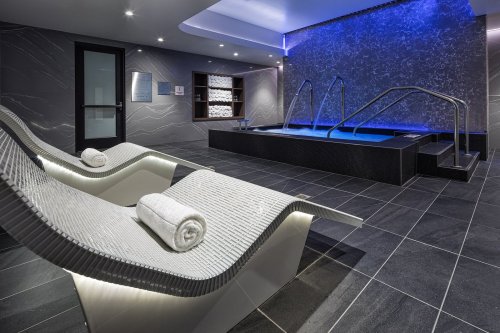 A modern spa area featuring lounge chairs, a hot tub, and ambient blue lighting on sleek tiled floors.