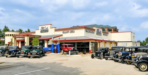 A vintage car garage with classic cars parked outside, featuring a craft beer sign and a retro architectural style.