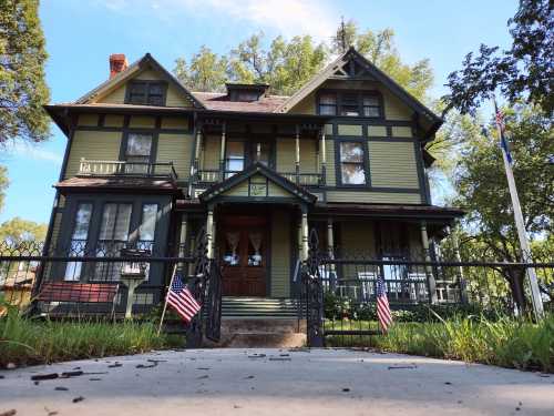 A large, historic green Victorian house with a porch, surrounded by a fence and small American flags at the entrance.